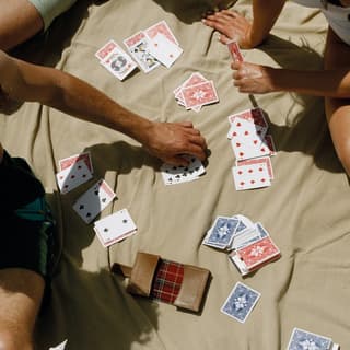 View from above of people playing games with red and blue decks of cards in the sun, lounging on a beige blanket.