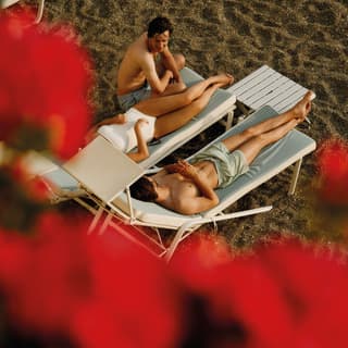 View down through red flowers to a woman in a white bathing costume and two men, relaxing and chatting on beach sun loungers.