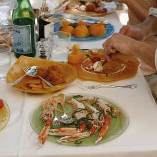 A plate of langoustines rests at the end of a shady lunch table where diners enjoy a meal with white wine and mineral water.