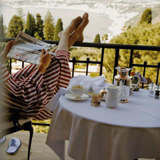 On a Grand Hotel Timeo balcony with sea view, a male guest sits next to a breakfast table with his feet up on the railings.