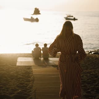 A woman in a robe walks up a wooden walkway, away from friends sitting by the gleaming shore in a hazy overexposed image.
