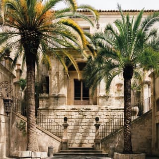 The cool paved courtyard of Palazzo Di Lorenzo del Castelluccio in Noto, Sicily, with palm trees and dual stone staircases.