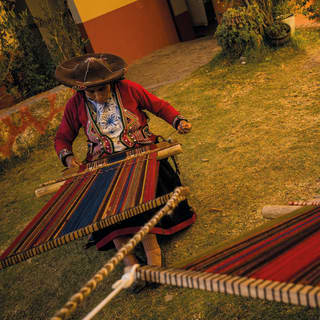 Two women in wide felt hats with handheld looms create traditional woven wool fabric in red, yellow, black and blue stripes.