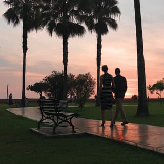 In rosy sunset light, a man and woman stroll arm-in-arm through the palm trees that line the Miraflores Boardwalk in Lima.