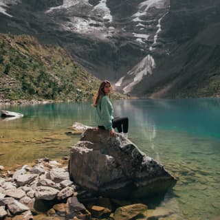 A young woman in poses on rock by the sapphire lagoon of Humantay Lake, cupped between snowy mountains in the highlands.