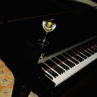 A classic chilled Gin Martini with olive garnish rests on the top of a black grand piano, seen at an angle from above.