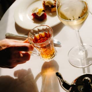 A guests holds a glass of amber tea at a table with Afternoon Tea delicacies and white wine at Botanica.