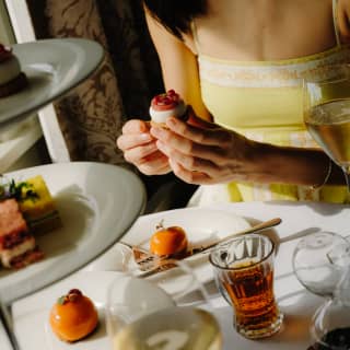 A woman in a yellow dress holds a cheesecake bite as she enjoys Afternoon Tea, with Apricot & Thyme Macarons and infusions.