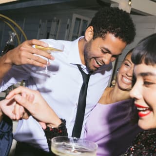 A man in a shirt and tie laughs with a woman in lilac and another in black sparkles as they share cocktails in coupe glasses.