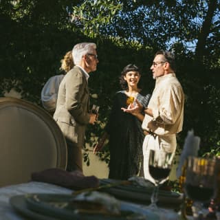 Two men and two women chat in the gardens of the Oak Tree Suite, standing in sunshine, viewed over a shaded dining table.