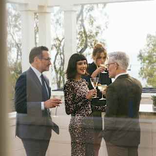 Three women in party dresses and two men in suits enjoy drinks on the Channel Islands Terrace, served by a barman in white.