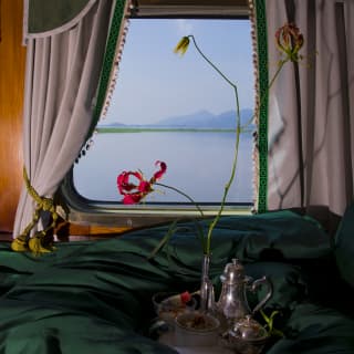 A tray with a silver tea set rests on a green duvet adorned with a flame lily that stands out against the misty window view.