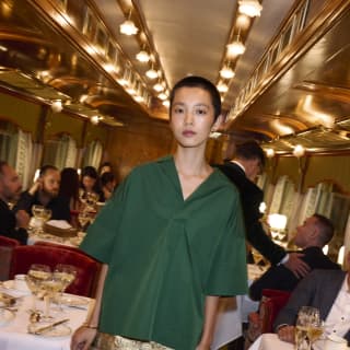 An elegant woman with short dark hair, in a green shirt and gold mini-skirt, walks through the busy, brightly-lit dining car.