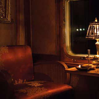 In a wood-panelled cabin, a table lamp picks up the sheen of a red silk seat by the window, which looks out onto dark night.