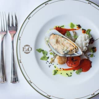 Looking down on a branded Eastern and Oriental Express dining plate with a cooked mussel starter presented with garnish.