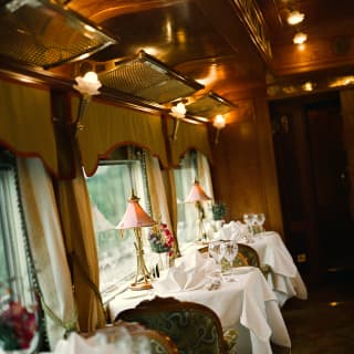 Tables in Adisorn Dining Car, with pink lamps, white tablecloths, wine glasses and fanned napkins, wait for dinner guests.