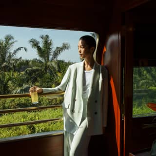 A woman with short dark hair in a loose, mint trouser suit stands on the part-shaded Observation Car deck, holding a drink.
