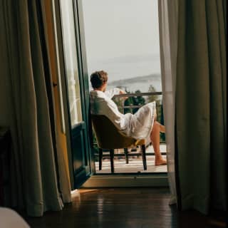A man in a white robe relaxes in a chair on a balcony, gazing at the views, seen from inside the suite through open doors.