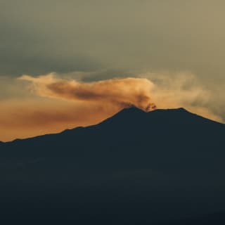 Dramatic image of smoke rising from the silhouetted Mount Etna, melting with the sunset to create plumes of orange and brown.