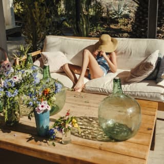 A girl sinks into the cream cushions of a large outdoor sofa, her whole head swallowed by a straw hat. Flowers fill the table