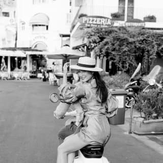 A young women holds onto her straw boater and smiles back at the camera as she rides pillion on a Piaggio scooter