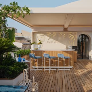 Three stools sit at an angle by the Louis Vuitton bar on a sun-dappled rooftop deck with lanterns, foliage and shade canopy.