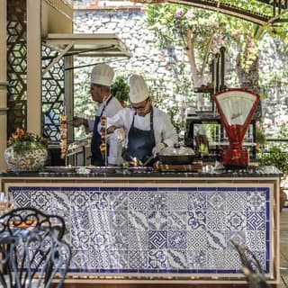 Blue majolica tiles adorn an outdoor kitchen. Two chefs in whites, toques and navy aprons work shaded by a bamboo awning