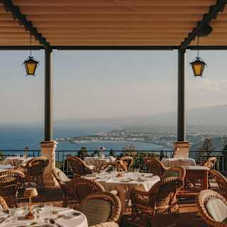 Laid tables with wicker chairs await guests on the Ristorante Timeo terrace, with a canopy and breathtaking coastline views.