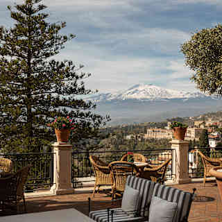 A large, furnished terrace framed by pines and olive trees overlooks the snow-dusted slopes of the cone-shaped Mount Etna