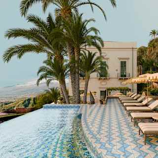 A row of loungers and parasols sit on Villa Timeo's blue and white-tiled terrace by an infinity pool with priceless views.