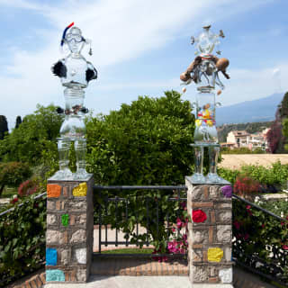 Two transparent statues decorated and raised on painted brick posts are the bright contribution of Marthine Tayou for MITICO.