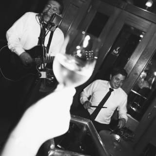 A guest raises a glass of white wine in soft-focus foreground as two musicians play behind, in an angled monochrome image.