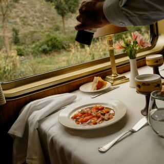 A waiter cracks pepper over a vibrant dish of Cured Trout with Nikkei sauce at a dining table with bread, and hillside views.