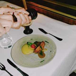 A waiter cracks pepper over a plate of pan-fried fish in Sudado sauce with puree, seen from above in a close-up dining detail.