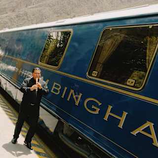 Angled image of a man in black as he plays the Peruvian flute at the edge of the platform by the blue Hiram Bingham train.