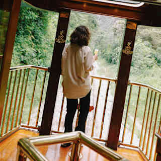 A woman in black trousers and khaki top stands by the rails of the Observation Car deck holding a drink, seen from behind.