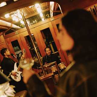 Angled image of a guitarist and drummer performing in the Bar Car as a woman, in soft-focus foreground, raises a drink.