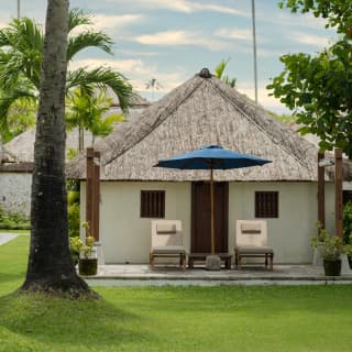 A parasol and sun chairs overlook a lush lawn outside a Balinese-style suite with white walls and triangular thatched roof.