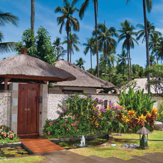 Thatched roofs and lofty palms hint at tropical luxury beyond the private entrance of a villa, bordered by exotic plants.