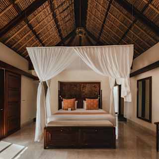 A high gabled ceiling with exposed beams rises above a four-posted bed with white drapes in the airy villa bedroom.