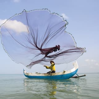 At sea, a fisherman in a hat and yellow top casts a circular net from his traditional white and blue Balinese outrigger boat.