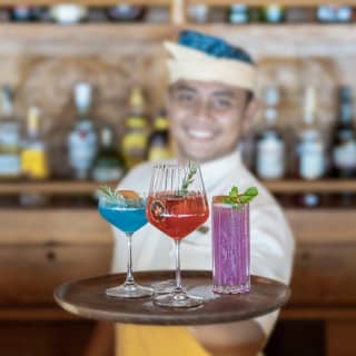 In soft focus, a smiling bartender extends a tray with three sharp-focus exotic cocktails in bright blue, deep red and lilac.
