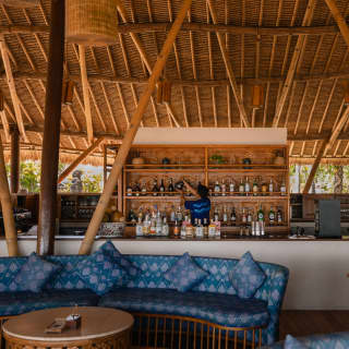 A thatched roof with giant supports towers above blue banquettes by the Puri Bar, where a barman is sorting the bottle shelf.