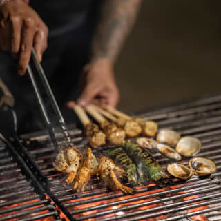 Details of a chef's hands using tongs to turn green tiger prawns, clams and fish sticks on the glowing embers of a hot grill.