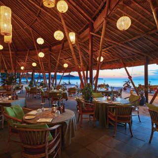 Late evening view across Nelayan restaurant, with tables laid and pendant lights glowing, to the sand and luminous sea.