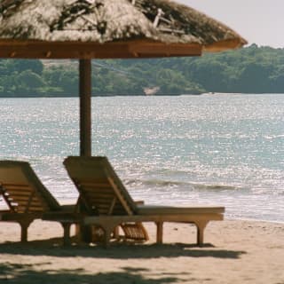 Two loungers and a thatched parasol sit by the glittering water on Jimbaran Beach with views to the land, south of the bay.