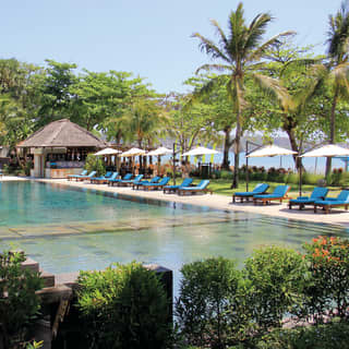 Clear, mirror-still outdoor pool surrounded by sunbeds, parasols and lush palms