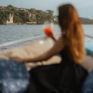 At the prow of the Dedari, in soft-focus, a long-haired woman in a black dress holds an orange drink and gazes at the coast.