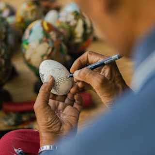 A local man decorates a painted white egg with a fin-tip pen, in an activity detail viewed over the artist's shoulder.