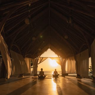 Two guests in lotus pose face the sea and sun, which bathes the yoga pavilion in honey light and shadows, seen from behind.